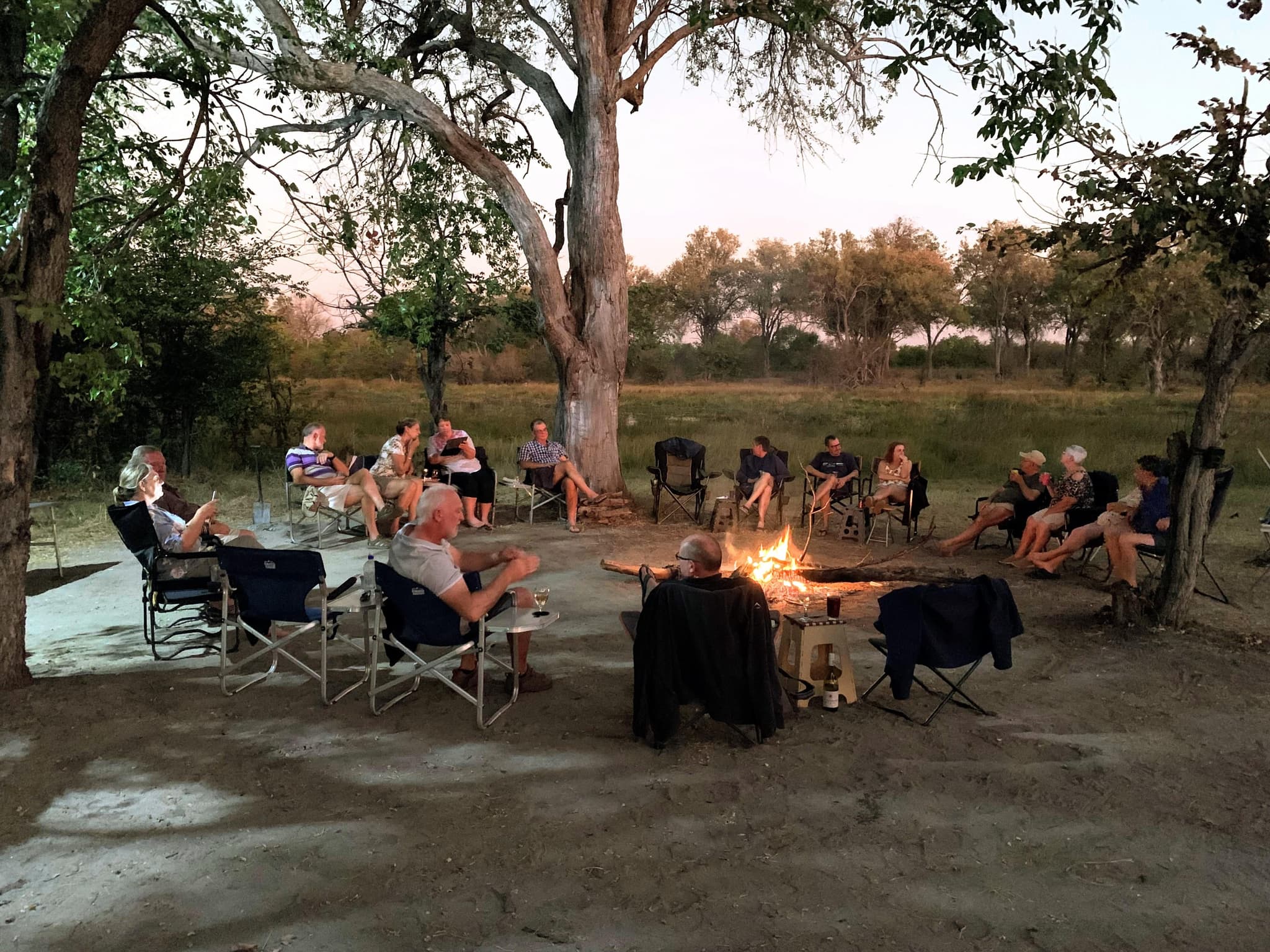 Safari travellers relaxing in camp chairs around a glowing campfire at dusk in the African bush